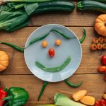 Fresh vegetables arranged creatively around a smiling plate on wooden table.